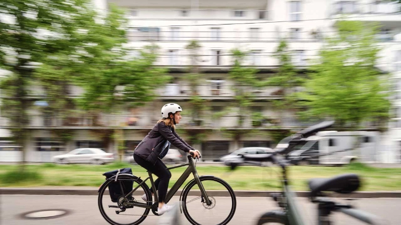Eine Radfahrerin in Bürokleidung fährt die Straße entlang, Sie hat ihre Tasche am Hinterradblech ihres Fahrrads befestigt