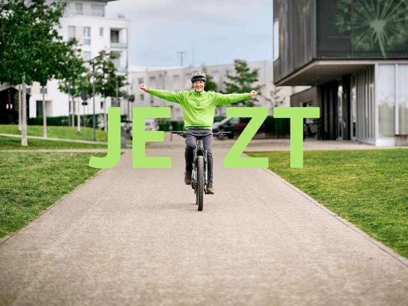 Radler fährt strahlend auf seinem Fahrrad mit gestreckten Armen mit Schriftzug "Jetzt"