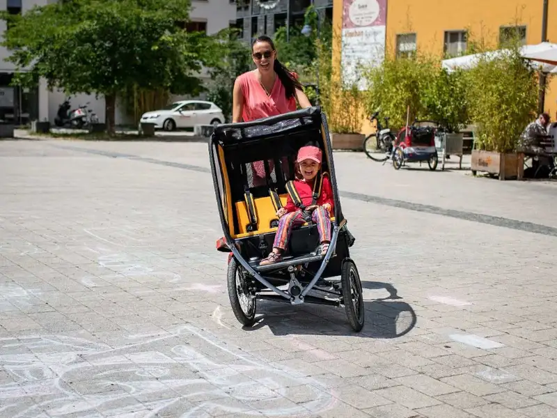 Eine Mutter fährt das Chike e-Bike in der Stadt. Ihr Kind sitzt angeschnallt darin. Beide lachen