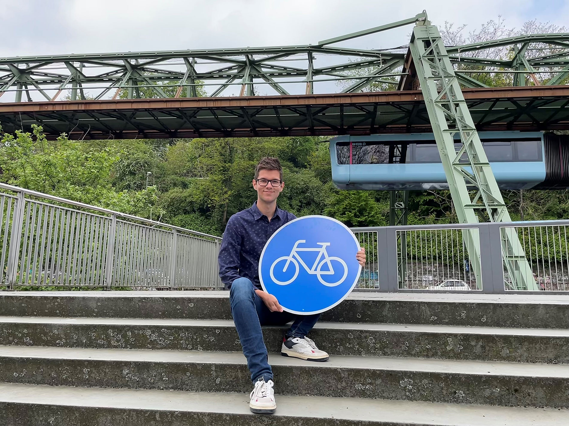 Leonard Arning sitzt mit Fahrradweg-Schild auf einer Treppe, im Hintergrund die Wuppertaler Schwebebahn